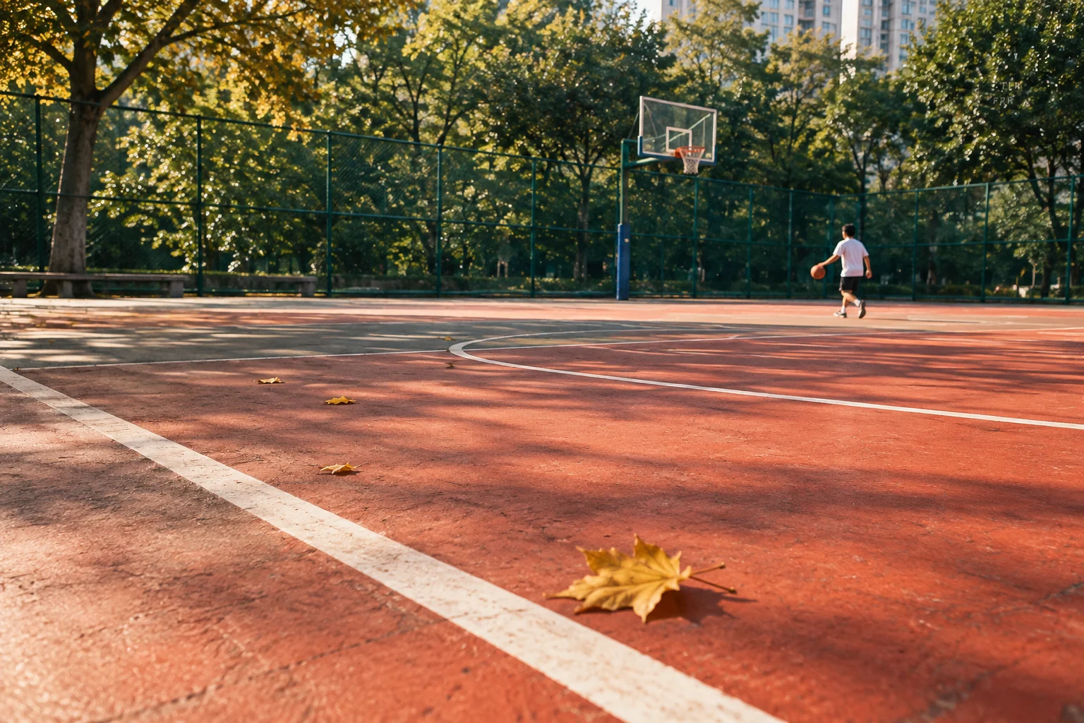 An orange basketball court, green fence, distant player, and yellow leaf.