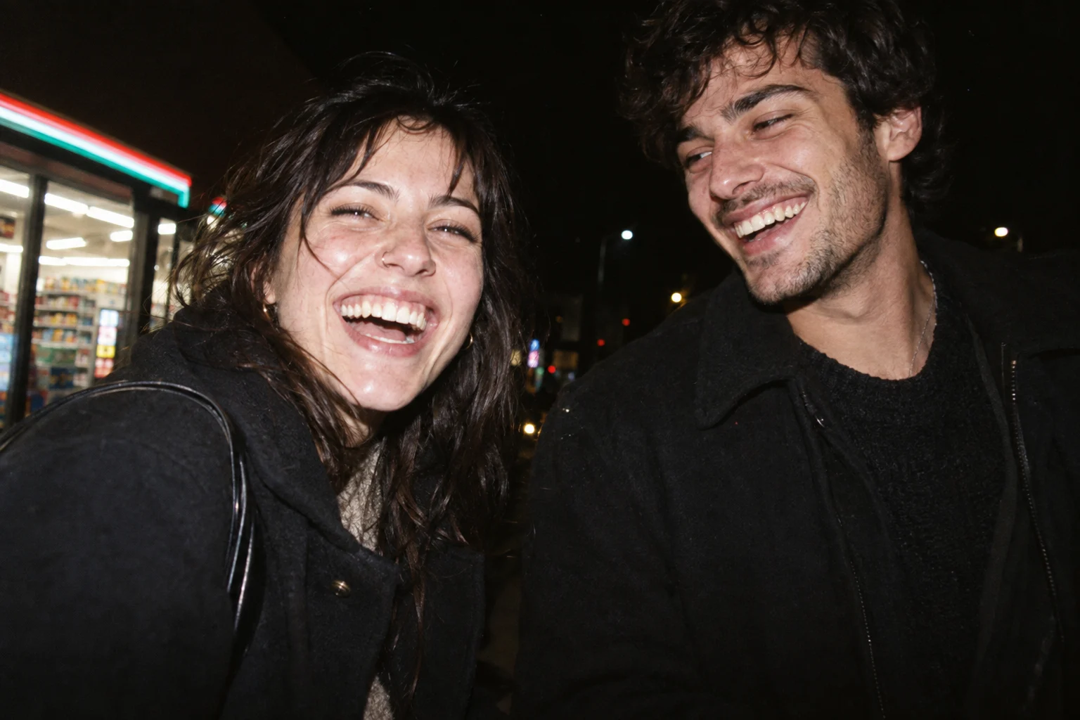 Two adult friends laughing outside at night, illuminated by direct camera flash.