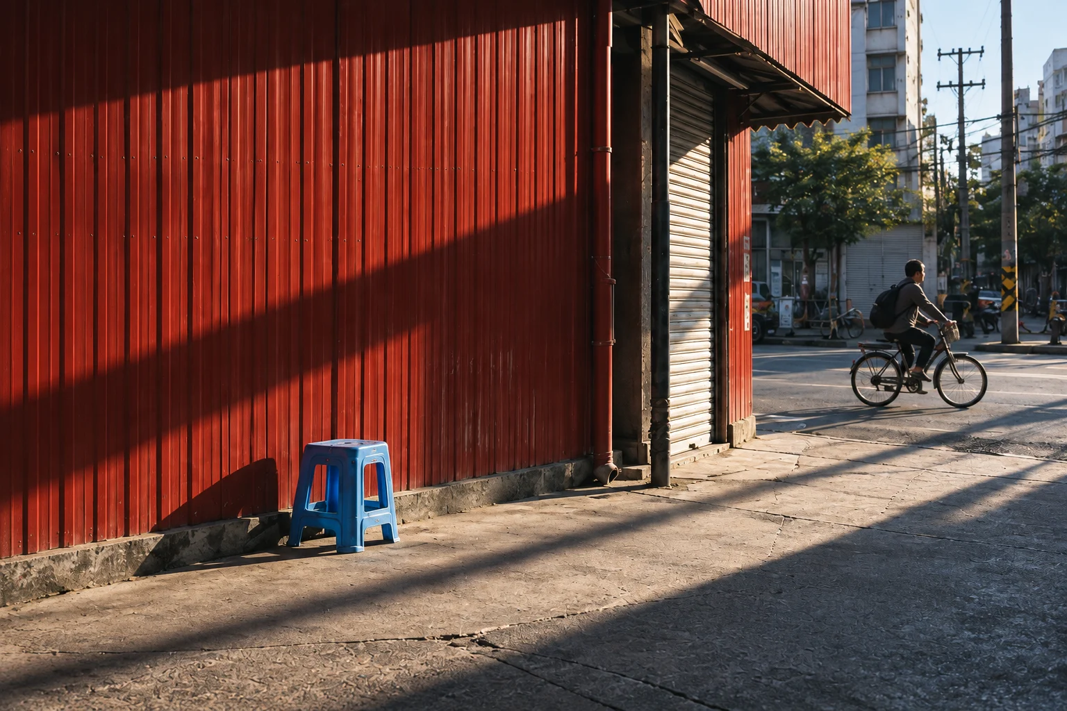 A cyclist passes a red wall and blue stool in sharp afternoon light.
