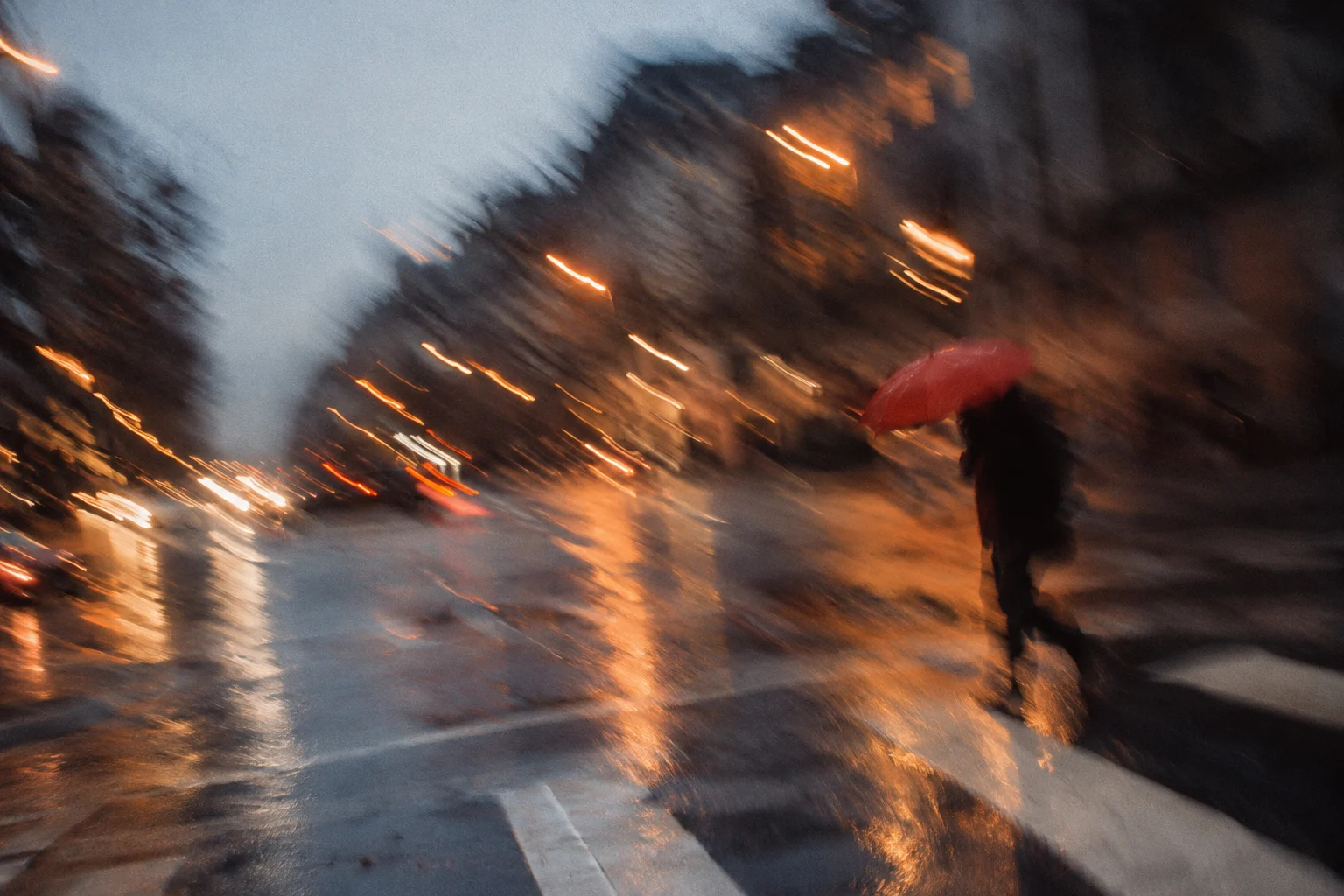 A red umbrella crosses a wet street with intentional slow-shutter motion blur.